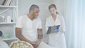 Mid-adult Caucasian woman talking with man sitting on bed in hospital ward and smiling. Portrait of professional doctor