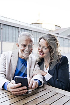 Mid adult couple looking at smartphone while sitting at outdoors table. Carefree senior man and woman using mobile phone