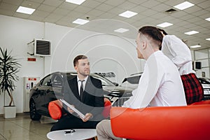 Mid-adult couple talking with sales agent and signing contract for new car in modern car showroom.