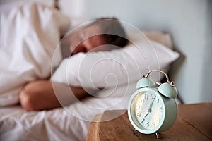 Mid adult man asleep in bed, alarm clock on the bedside table in the foreground, focus on foreground