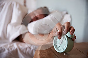 Mid adult man asleep in bed, reaching out to alarm clock on the bedside table in the foreground