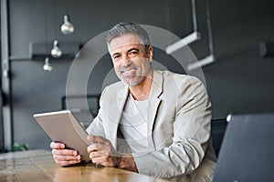 Mid aged business man holding digital tablet sitting in modern office. Portrait