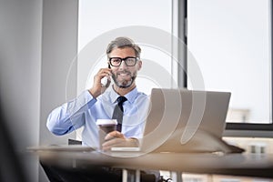 Mid aged businessman sitting at office desk and using cellphone and laptop