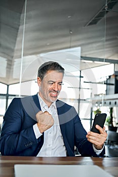 Mid aged happy business man looking at cell phone celebrating success in office.