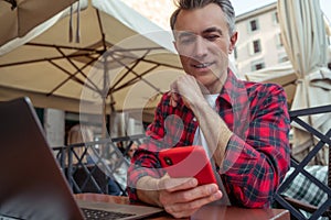 Mid aged man in plaid shirt sitting at the table in the street cafe with a phone in hands