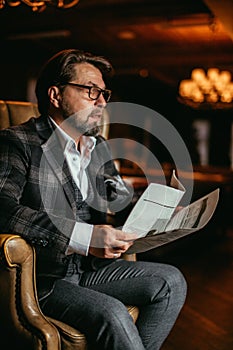 Mid-aged well-dressed man sitting in arm-chair in living room reads newspaper