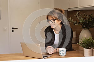 Mid aged woman standing at the kitchen at home and using laptop