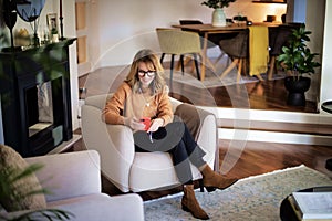 Mid aged woman using earphones and cellphone and sitting in an armchair at home