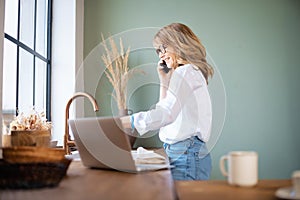 Mid aged woman using mobile phone and laptop in the kitchen at home