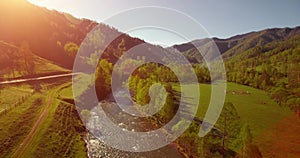 Mid air flight over fresh mountain river and meadow at sunny summer morning. Rural dirt road below.