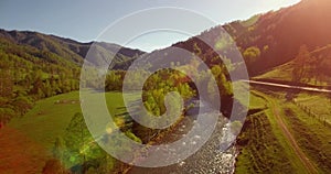 Mid air flight over fresh mountain river and meadow at sunny summer morning. Rural dirt road below.