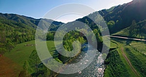 Mid air flight over fresh mountain river and meadow at sunny summer morning. Rural dirt road below.