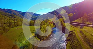 Mid air flight over fresh mountain river and meadow at sunny summer morning. Rural dirt road below.