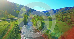 Mid air flight over fresh mountain river and meadow at sunny summer morning. Rural dirt road below.