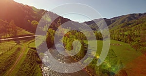 Mid air flight over fresh mountain river and meadow at sunny summer morning. Rural dirt road below.