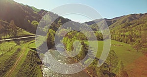 Mid air flight over fresh mountain river and meadow at sunny summer morning. Rural dirt road below.