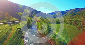 Mid air flight over fresh mountain river and meadow at sunny summer morning. Rural dirt road below.