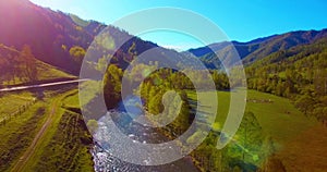 Mid air flight over fresh mountain river and meadow at sunny summer morning. Rural dirt road below.