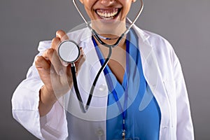 Mid section of african american female doctor holding stethoscope smiling against grey background