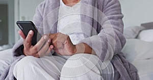 Mid section of african american senior woman using smartphone while sitting on the bed at home