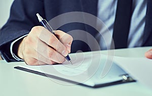 Mid section of businessman in suit filling and signing with blue pen partnership agreement form clipped to pad closeup.