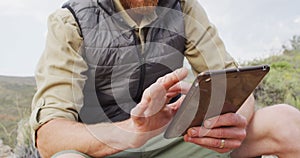 Mid section of caucasian male survivalist sitting on mountain, using tablet