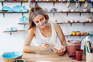 Mid-shot of potter decorating clay mug after firing in oven. Woman in white tanktop enopying creative procces of pottery coloring