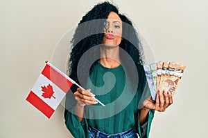Middle age african american woman holding canada flag and dollars looking at the camera blowing a kiss being lovely and sexy