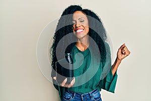 Middle age african american woman holding oil motor bottle screaming proud, celebrating victory and success very excited with