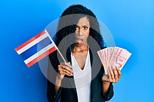 Middle age african american woman holding thailand flag and baht banknotes in shock face, looking skeptical and sarcastic,