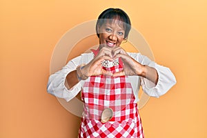 Middle age african american woman wearing baker uniform smiling in love doing heart symbol shape with hands