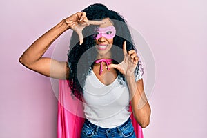 Middle age african american woman wearing super hero costume smiling making frame with hands and fingers with happy face