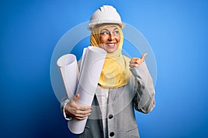 Middle age architect woman wearing muslim hijab and security helmet holding blueprints pointing and showing with thumb up to the