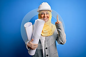 Middle age architect woman wearing muslim hijab and security helmet holding blueprints surprised with an idea or question pointing