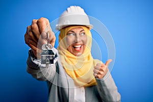 Middle age architect woman wearing muslim hijab and security helmet holding home keys pointing and showing with thumb up to the