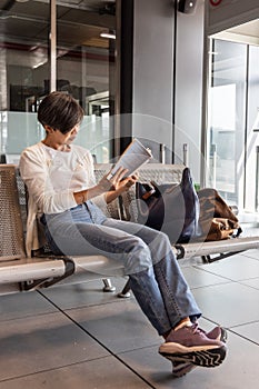 MIddle age Asian woman with luggage reading book while waiting for departure in airport terminal