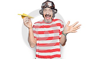 Middle age aviator man wearing vintage helmet and glasses holding paper airplane celebrating achievement with happy smile and