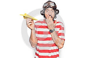 Middle age aviator man wearing vintage helmet and glasses holding paper airplane covering mouth with hand, shocked and afraid for