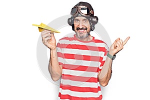 Middle age aviator man wearing vintage helmet and glasses holding paper airplane smiling happy pointing with hand and finger to