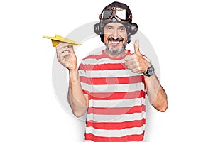 Middle age aviator man wearing vintage helmet and glasses holding paper airplane smiling happy and positive, thumb up doing