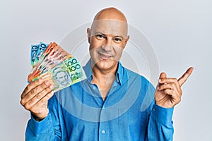 Middle age bald man holding australian dollars smiling happy pointing with hand and finger to the side