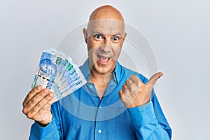 Middle age bald man holding south african 100 rand banknotes pointing thumb up to the side smiling happy with open mouth