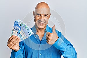 Middle age bald man holding south african 100 rand banknotes smiling happy and positive, thumb up doing excellent and approval
