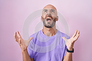 Middle age bald man standing over pink background crazy and mad shouting and yelling with aggressive expression and arms raised