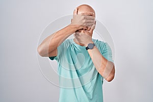 Middle age bald man standing over white background covering eyes and mouth with hands, surprised and shocked