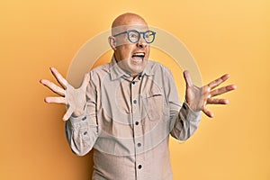 Middle age bald man wearing casual clothes and glasses crazy and mad shouting and yelling with aggressive expression and arms