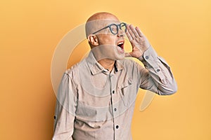 Middle age bald man wearing casual clothes and glasses shouting and screaming loud to side with hand on mouth