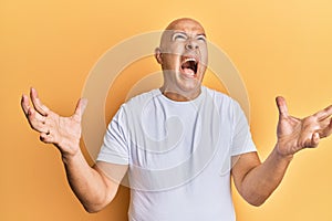 Middle age bald man wearing casual white tshirt crazy and mad shouting and yelling with aggressive expression and arms raised