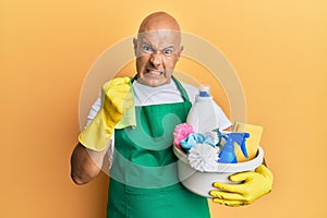 Middle age bald man wearing cleaner apron holding cleaning products annoyed and frustrated shouting with anger, yelling crazy with