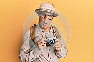 Middle age bald man wearing explorer hat and vintage camera in shock face, looking skeptical and sarcastic, surprised with open